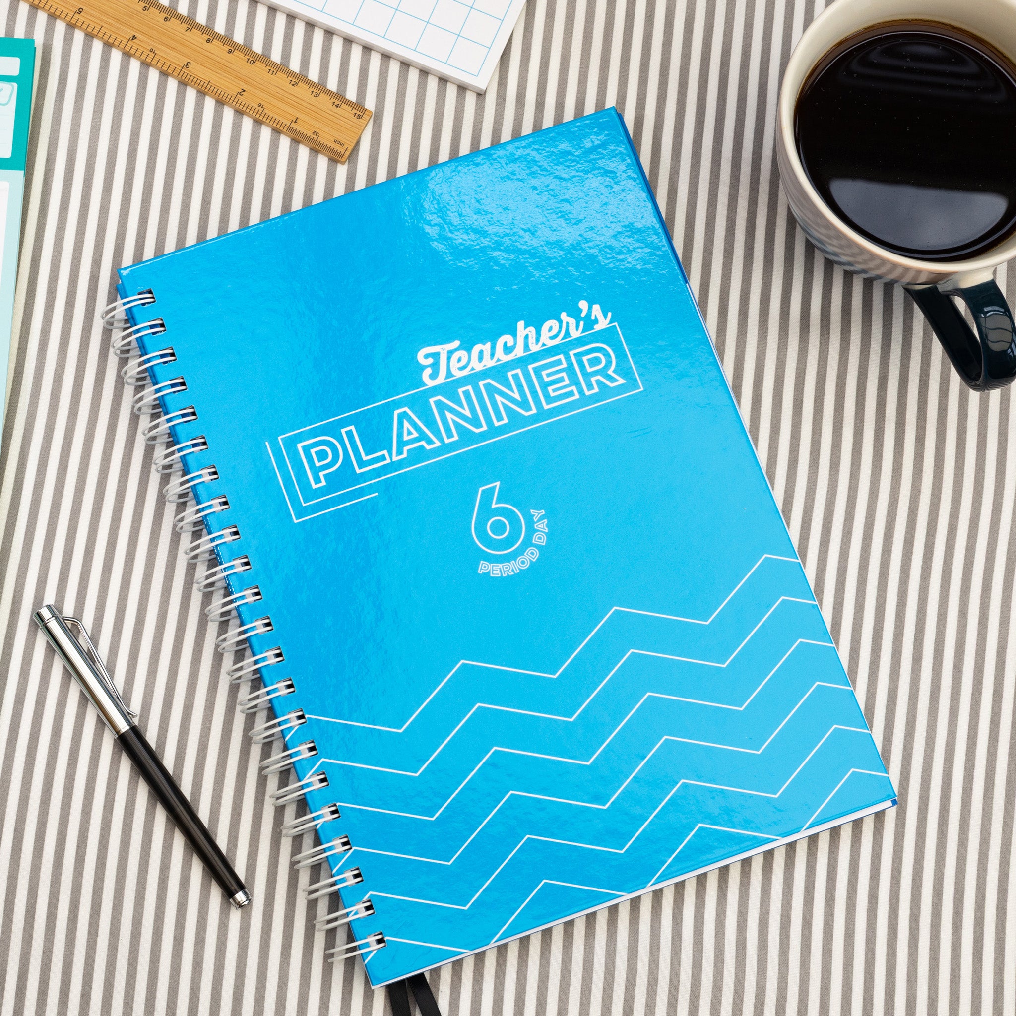 Silvines Blue Teacher's Planner Book On A Striped Table Cloth With A Pen & Coffee Cup.