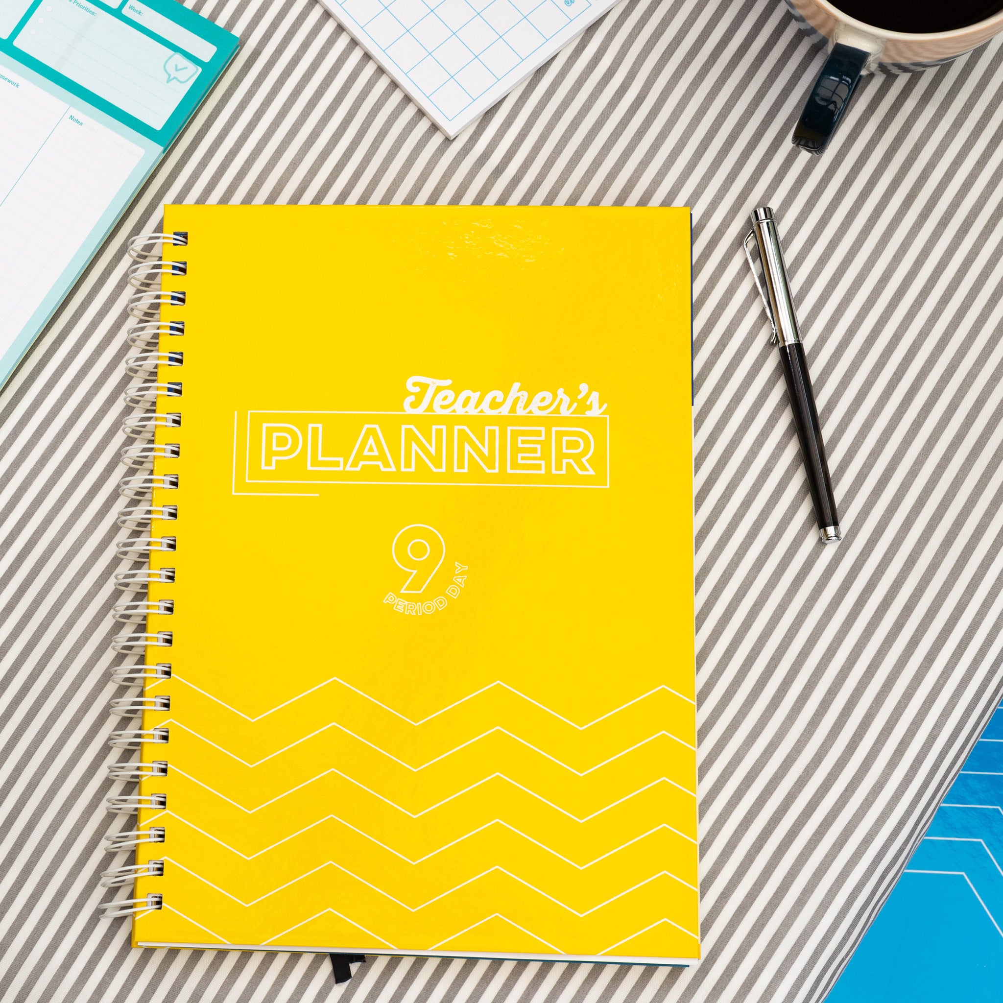 Silvines Yellow 9 Period Teacher's Planner On A Striped Table Cloth With A Pen & Coffee Cup.