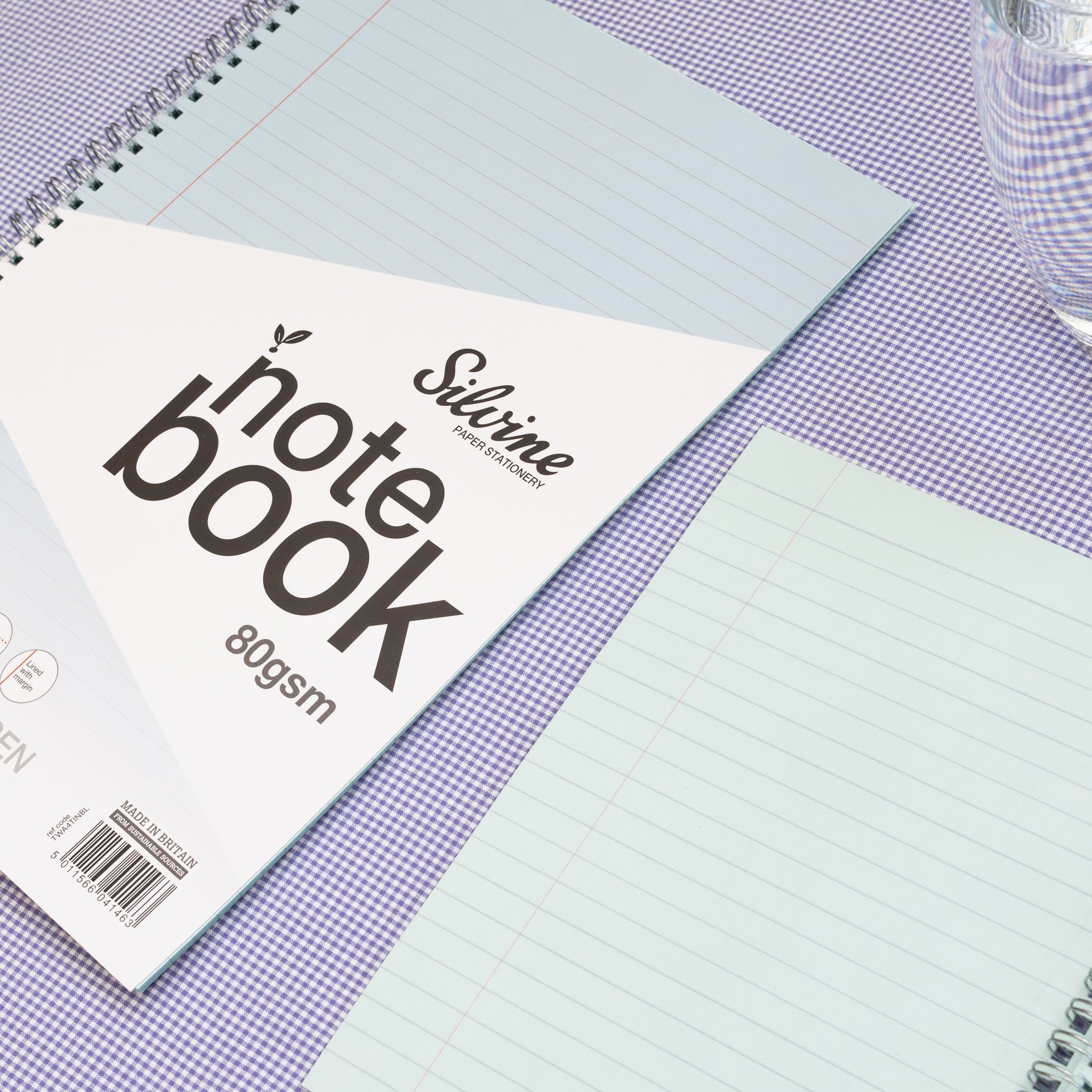 Two Silvine Blue Tinted Paper Notebooks, One Closed & One Open Displaying The Lined Paper On A Purple Gingham Background